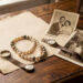 Bague ancienne, collier de perles et montre vintage disposés sur un bureau en bois près de papier ancien vierge et de photographies de famille sépia, baignés d’une lumière douce.